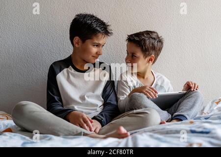 Smiling brothers in sleepwear sitting on cozy bed and watching movie together while entertaining at home Stock Photo