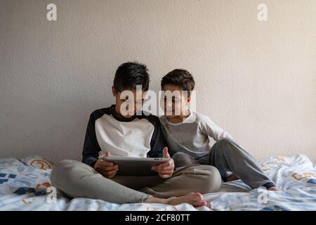 Smiling brothers in sleepwear sitting on cozy bed and watching movie together while entertaining at home Stock Photo