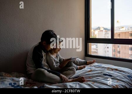 Smiling brothers in sleepwear sitting on cozy bed and watching movie together while entertaining at home Stock Photo