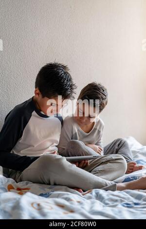 Brothers in sleepwear sitting on cozy bed and watching movie together while entertaining at home Stock Photo