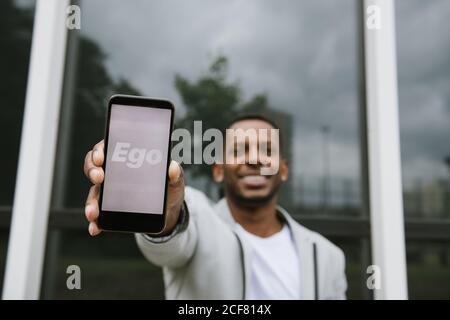 Stylish cheerful African American man holding smartphone with Ego text on screen on background of reflecting wall Stock Photo
