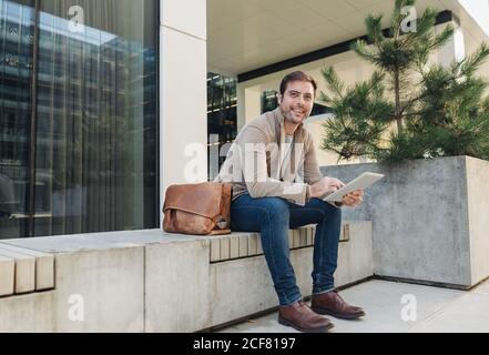man manager smiling and browsing on a tablet while sitting outside contemporary building on city street Stock Photo