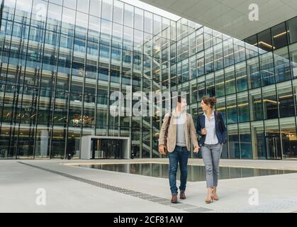 Full body delighted colleagues smiling and looking at each other while walking outside contemporary building after work Stock Photo
