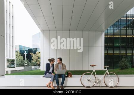 Delighted businesspeople smiling and browsing laptop together while sitting outside modern building near bicycle on city street Stock Photo