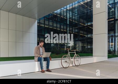 Full body happy man manager smiling and browsing laptop while sitting outside near bicycle on a contemporary building on city street Stock Photo