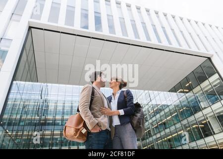 From below happy couple coworkers kissing each other while standing outside modern building on city street after work Stock Photo