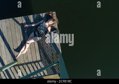 Top view of beautiful Woman in black swimsuit and hat lying down on wooden pier of lake on clear blue sky and forest background Stock Photo