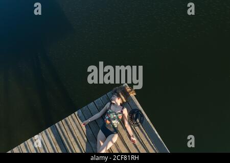 Top view of beautiful Woman in black swimsuit and hat lying down on wooden pier of lake on clear blue sky and forest background Stock Photo