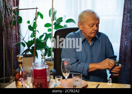 Serious casual aged male in blue shirt opening bottle of champagne while sitting at table during family Christmas dinner at home Stock Photo