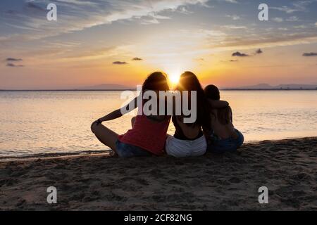 Back view of bronzed female friends hugging and relaxing on beach at sunset in summer in back lit Stock Photo