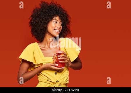 Cheerful young African American lady with curly hair holding red jar with straw and enjoying beverage on red background Stock Photo