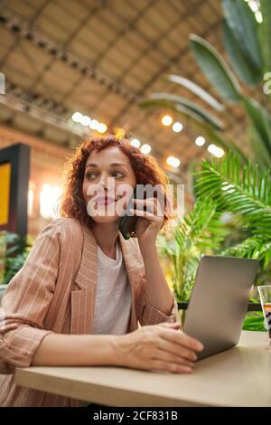 Cheerful young Woman working on laptop in cafe and talking phone Stock Photo