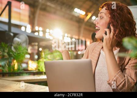 Red haired excited young female sitting at table in cafe speaking on phone while working on laptop and pointing away Stock Photo