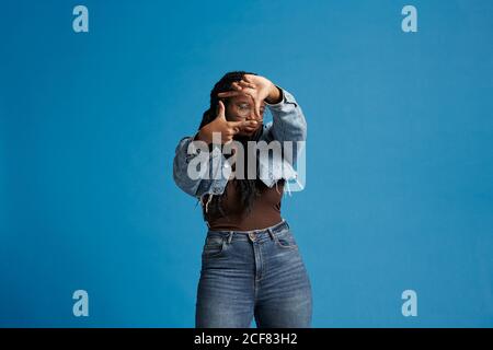 Happy black female with braids in denim jacket smiling and looking at camera while framing face with hands against blue background Stock Photo