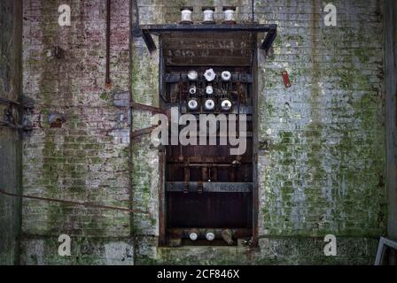Old broken rusty mechanism with worn steel pipes of coils and rods in dilapidated gray brick wall covered with moss in abandoned factory Stock Photo