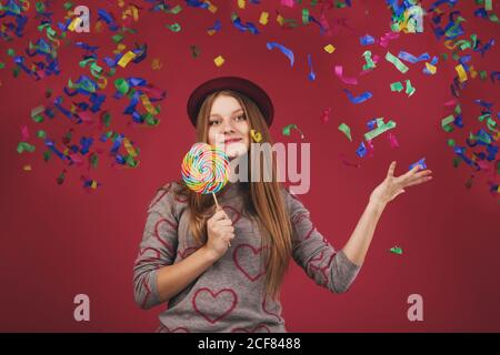 Portrait of a teen girl throws up a multi-colored tinsel and confetti. beautiful girl wearing hat holding big striped lollipop Stock Photo