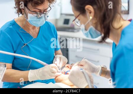 Confident specialists in surgical gloves putting seal and working with dental tools in mouth of patient lying in chair in cabinet Stock Photo