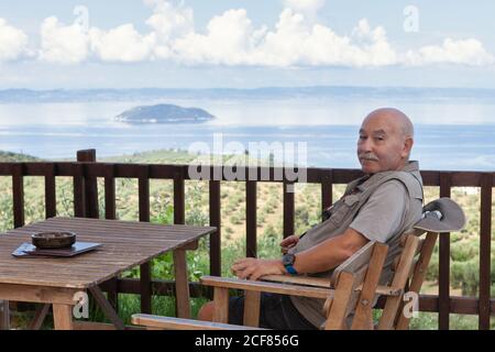 Side view of content retired man with sitting in chair at table on terrace with beautiful ocean view on background, Greece Stock Photo