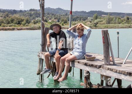 Relaxing elderly man and Woman in hats and sunglasses enjoying time together while sitting on pier a in Halkidiki, Greece Stock Photo