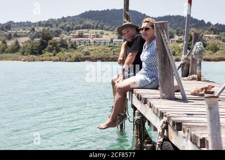 Relaxing elderly man and Woman in hats and sunglasses enjoying time together while sitting on pier and looking at camera in Halkidiki, Greece Stock Photo