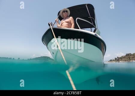 From below elderly man in hat and sunglasses sailing on black and white boat in calm turquoise water of Halkidiki, Greece Stock Photo