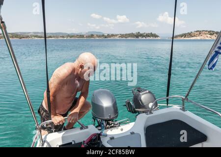 Tanned elderly man climbing on yacht holding rail of boat while swimming in turquoise water, Halkidiki, Greece Stock Photo