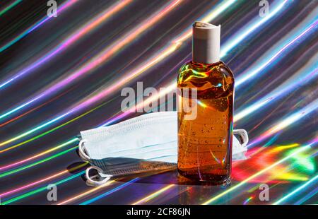 Protective medical face mask and bottle of brown sanitizer gel on holographic shiny background Stock Photo