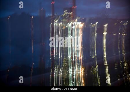 Blurred night white yellow lights of brightly lighting area of city with industrial pipes at dusk on cloudy evening Stock Photo