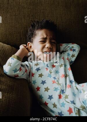 Cute African American boy in pajamas lying on comfortable sofa and starting to cry at home Stock Photo