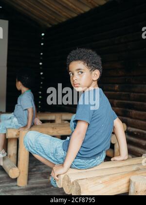 Cute African American boy in casual outfit looking over shoulder while sitting on wooden structure inside lumber building Stock Photo