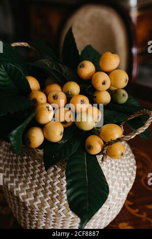 Wicker basket with loquat fruits and green leaves placed on wooden table in vintage styled fashioned interior of retro dining room Stock Photo