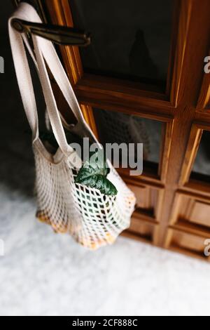 Reusable white string bag with fresh loquat fruits on branches hanging on knob of wooden door inside room with dark light Stock Photo