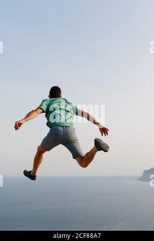 Low angle back view of unrecognizable male in casual clothes jumping high against blue sky and sea during summer evening Stock Photo