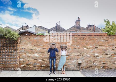 Couple in casual wear standing near brick wall in city and putting on wooden fruit boxes on heads while behaving ridiculously Stock Photo