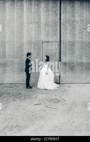 Black and white groom in classy tuxedo and bride in white wedding dress standing on background of shabby building during wedding day Stock Photo