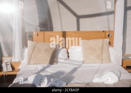 Made-up bed with white linen and beige pillows under transparent roof on sunny day Stock Photo