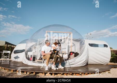 Stylish Woman sitting on boyfriend laps and looking through telescope at sky near bubble hotel Stock Photo