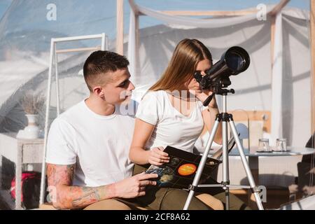 Stylish Woman sitting on boyfriend laps and looking through telescope at sky near bubble hotel Stock Photo