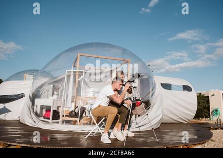 Stylish Woman sitting on boyfriend laps and looking through telescope at sky near bubble hotel Stock Photo