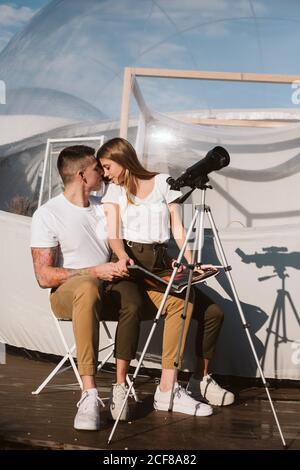 Stylish Woman sitting on boyfriend laps , reading and and looking through telescope at sky near bubble hotel Stock Photo