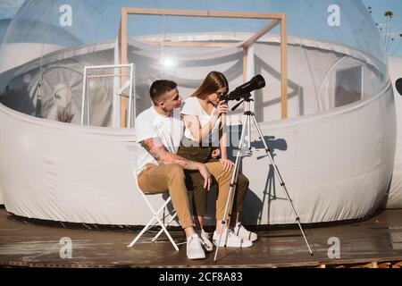 Stylish Woman sitting on boyfriend laps and looking through telescope at sky near bubble hotel Stock Photo