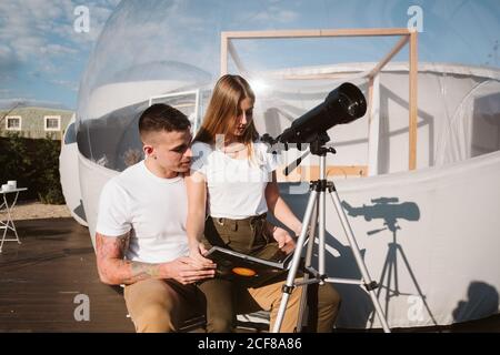 Stylish Woman sitting on boyfriend laps , reading and and looking through telescope at sky near bubble hotel Stock Photo