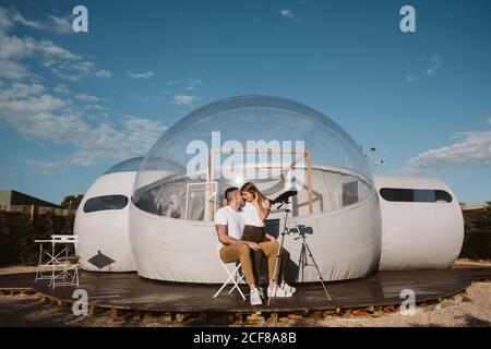 Stylish Woman sitting on boyfriend laps and looking through telescope at sky near bubble hotel Stock Photo