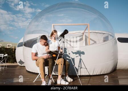 Stylish Woman sitting on boyfriend laps , reading and and looking through telescope at sky near bubble hotel Stock Photo