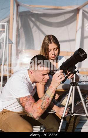 Stylish Woman sitting on boyfriend laps and looking through telescope at sky near bubble hotel Stock Photo