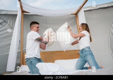 Cheerful young couple having fun during pillow fight on bed in big tent with transparent roof Stock Photo