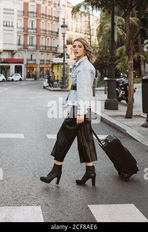 Side view of stylish young Woman with suitcase smiling and looking away while crossing city street Stock Photo