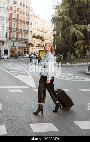 Side view of stylish young Woman with suitcase smiling and looking away while crossing city street Stock Photo
