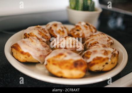 Closeup of tasty and appetizing homemade cinnamon rolls with sweet icing topping placed on white round dish on black surface in kitchen at home Stock Photo