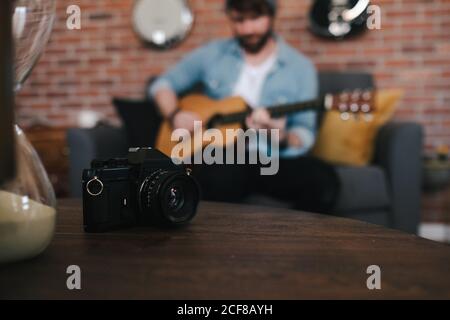 Crop bearded guy in denim jacket playing acoustic guitar while sitting on cozy couch in front of wooden table with photo camera and hourglass at home Stock Photo